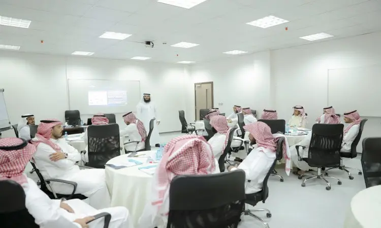 يجتمع عدد من الرجال مرتدين الغترة السعودية أو الشماغ مع العقال، بالإضافة إلى البشت السعودي التقليدي، حول طاولات في قاعة حديثة وواسعة داخل قاعة الألعاب في جامعة القصيم، مشاركة في دورة مخصصة للمعلمين، بينما يجتمع المحاضر أمامهم بقرب شاشة عرض لي أطراف الراحة.