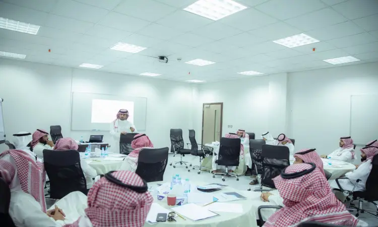 يجلس مجموعة من الرجال مرتدين الغترة أو الشماغ السعودي مع العقال والبشت السعودي، حول طاولات مستديرة في القاعة مضيئة في القصيم، حيث يصغون باهتمام لمقدمة العرض يناقش برنامج تطوير لكن بالقرب من شاشة عرض وسبورة بيضاء.