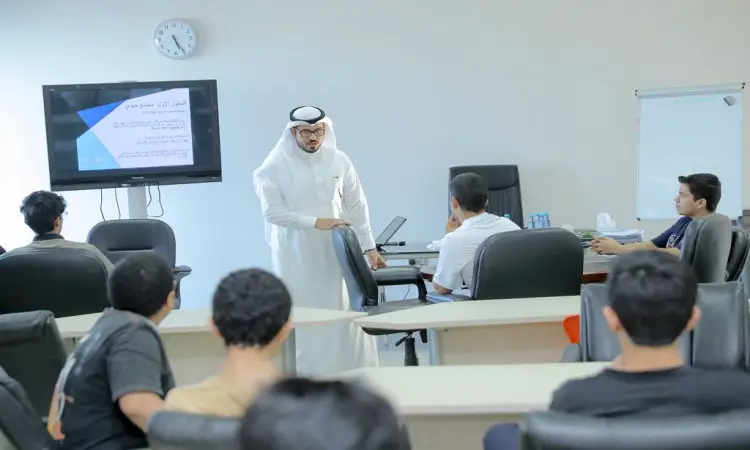 رجل يرتدي غترة سعودية وعقالًا وبشتًا رسميًا في قاعة الدعوة لحضور القصيم، وعمًا عرضًا أكاديميًا من الطلاب الجالسين. يناقش من خلال عرض دور الشباب، وذلك من خلال عرض النصوص المعروضة باللغة العربية، ويتناول في راديو ساعة الحائط وسبورة بيضاء المساهمين في إبراز كافة المشرق والرسمية.