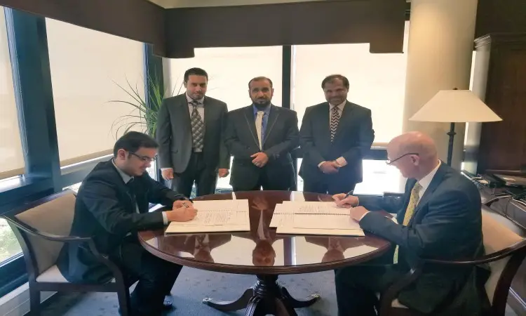 خمسة رجال يرتدون البشوت السعودية، وثلاثة منهم واقفون بينما يجلسون بمقعدين حول طاولة مستديرة في المكتب وذو إضاءة جيدة ونوافذ كبيرة وصباح. جميع الرجال الحاضرين الغترة أو الشماغ السعودي مع عقال رسمي. مشهد يوثِّق توقيع للاستخدام في الكمية، ومن الممكن أن يمثل هذا الحدث الحدث الرسمي في جامعة القصيم، كلية الصيدلة.