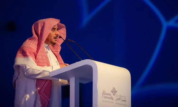 A distinguished gentleman dressed in traditional Saudi dress - a white ghutra, black headband, and an elegant Saudi bisht - delivers a speech from behind the podium. He stands in front of a blue background, addressing the audience through a microphone. The official logo and name of Qassim University in Arabic and English are projected on the stage during the closing ceremony of the university's summer club (closing ceremony of the university's summer club).