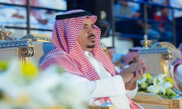 A man dressed in the authentic Saudi uniform of a Saudi Ghutrah, a Saudi Qatara, and a Saudi Besht over his white ghana sits in a plush chair and applauds as the Qassim University Club's summer culmination and commissioning is underway.