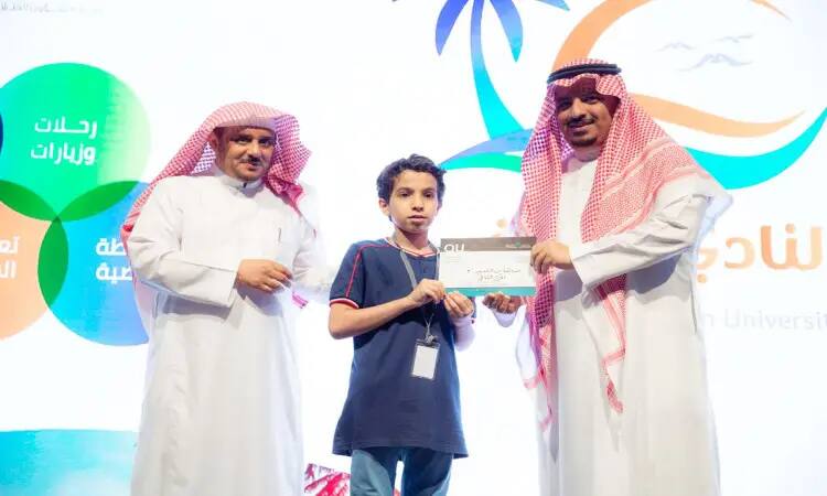 A young student receives a certificate of appreciation from two men dressed in a ghutra and aqal, and the official Saudi bisht, on a brightly lit stage during the conclusion of a summer club in Qassim. Consider creating colorful designs and bold text in Arabic to enliven the summer club.