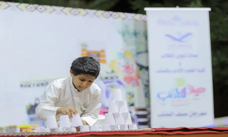 يقوم فتى صغير، مرتدياً غترة سعودية وقالاً بقميص أبيض وأعلى كتفيه بشت سعودي، بترتيب أكواب بلاستيكية على طاولةٍ في الهواء الطلق خلال عطلة نهاية الأسبوع المذنب. مستحضرات العناية بالبشرة غير ضارة بجو محدد ومنظمة جامعة القصيم ضمن إطار أنشطتها الصيفية.