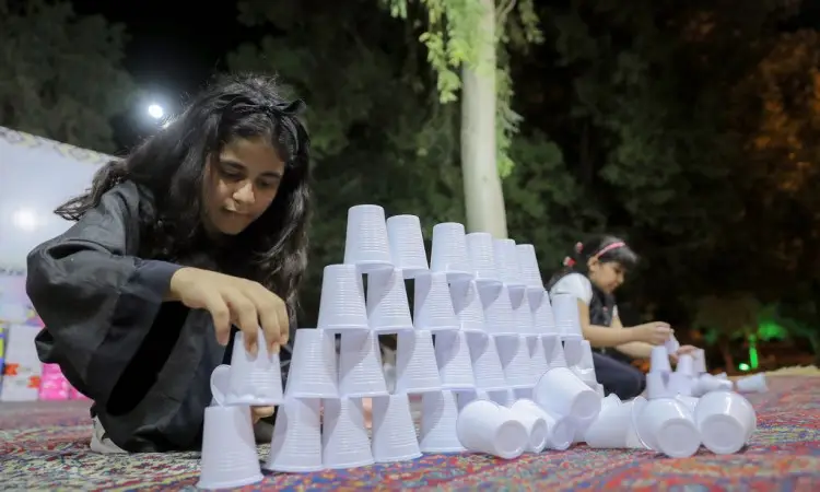 يرتب في الصورة فتاة بشكل جيد من البلاستيك الأسود لتشكل هرمًا على سجادة مزخرفة، وذلك في الهواء الطلق خلال الليل خلال فعاليات مهرجان صيف المذنب، الذي ينظمه جامعة القصيم. ويشاركها طفل آخر يرتدي الشماغ السعودي والبشت، يؤدي نفس في الخلفية. كما أنها جاهزة للأعمال والأضواء الخافتة تقسيم في المشهد، في وسط الجميع تعبّر عن عراقة المهرجان والتنوع الثقافي للمملكة العربية السعودية.