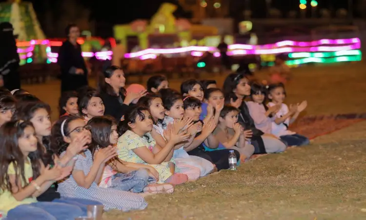 ضبط مجموعةٌ من الأطفال على العشب في المساء، يصفقون بحماس ويتابعون بعض فعاليات مهرجان صيف المذنب. تضيء الأنوار الملونة أرجاء المكان، مضفية جواً موضوعاً ومبهجاً. ملحوظه الأطفال بملابسهم التقليدية، حيث ظهرت الفرق الغترة أو الشماغ والعقال، بينما ارتدى بعض البالغين البشت السعودي في خطاب يعكس الأصالة وعمق الانتماء الوطني. احترام ملامح الأطفال حماسهم وتفاعلهم مع الانضمام وسط تسودها البهجة والروح المجتمعية التي ترعاها جامعة القصيم.