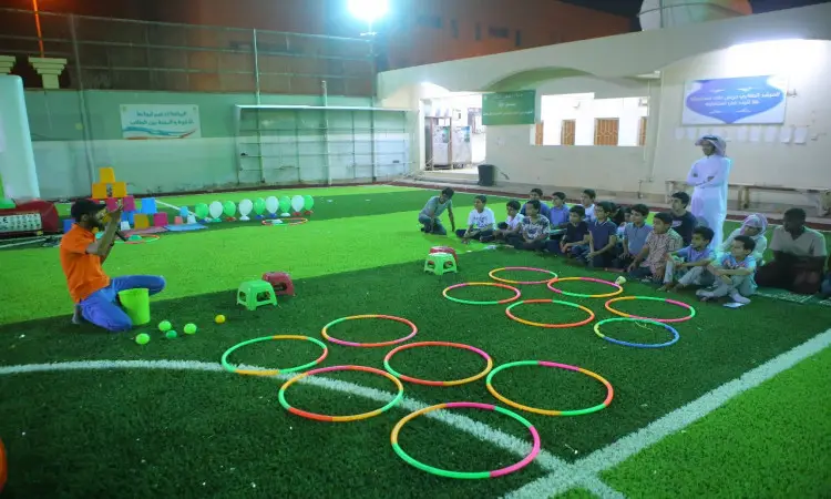 يجتمعون مجموعة من الأطفال على البلاستيك الصناعي، يراقبون مدربًا يتواصلون بجوار أطواق الهولا والملصقات البلاستيكية الملونة؛ فيما يتعلق بأحد فقط من برنامج ليالي عمادة خدمة المجتمع مرتديًا الغترة والعقال والبشت السعودي على مقربةٍ من ساحة الشؤون الخارجية المسائية، حيث تقضي البالونات وألعاب المساحات في المساحات.