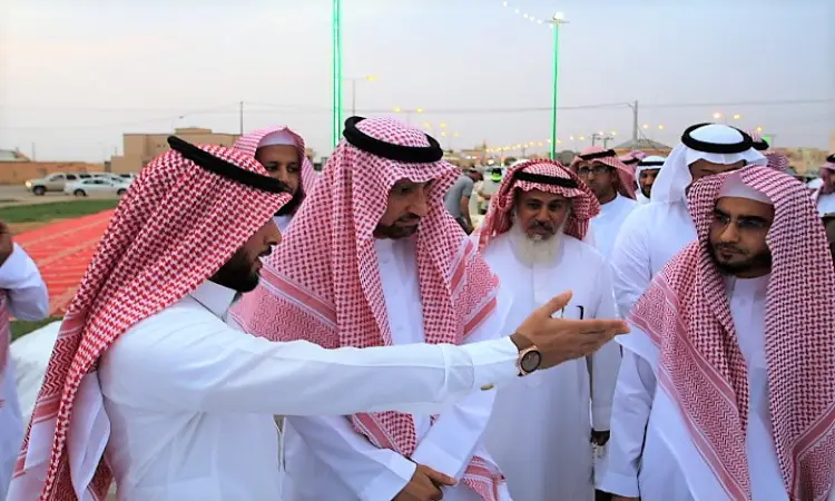 يجتمع مجموعة من الرجال مرتدين الزي السعودي الأصلي، والذي يشمل الثوب الأبيض، والشماغ أو الغترة السعودية مع العقال، بالإضافة إلى البشت السعودي، وذلك في الهواء الطلق من خلال فعاليات برنامج ليالي عمادة خدمة المجتمع في القصيم. وهي تشير إلى أثناء حديثه، بينما تبدو في الخلفية ضوء آخر من الحضور.