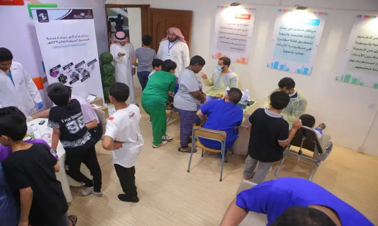 في قاعة عيادة مضيئة في جامعة القصيم، يتفاعل الأطفال ومنسوبو الرعاية الصحية—يرتدي الشركاء الغترة أو الشماغ والعقال والبشت السعودي أو الزي الطبي والكمامات—ضمن فعاليات خدمة المجتمع. وتزين الملصقات توعوية، مما يضفي على المكان المناسب ترحيبية تليق بكل مستفيد.