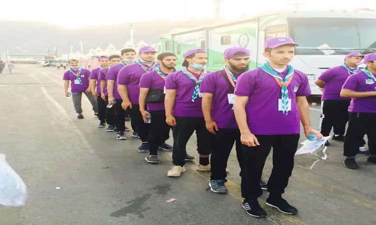 A group of Qassim University Jawalat youth are lined up in a fixed line outdoors, wearing uniform purple shirts, wearing a shemagh or Saudi ghutra with an aqal, and wearing the Saudi bisht on their shoulders. A number of them also bring blue necks and carry water while performing their duties as a service to the pilgrims.
