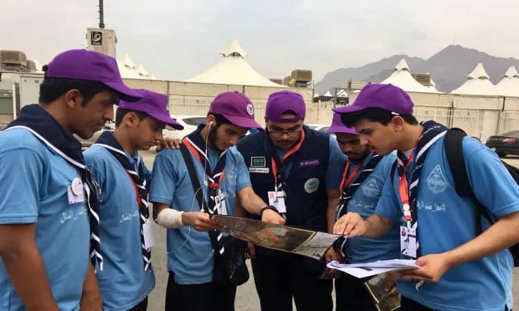 A team of Qassim University Rangers gathered in their blue uniforms, complete with a shemagh or Saudi ghutra with an aqal, and the leader wearing a Saudi besht over his shoulders. They were all in a square contemplating the free map next to the team's crew, sharing with them the white tents and the slab of mountains on the distant horizon.