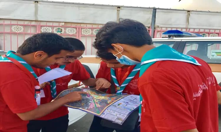 Four Qassim University Jawala youth, dressed in red shirts with a Saudi shemagh, or Saudi ghutra, headband and bisht, check a reading in the open air. A presence in a police car and a tent as part of their support activities to serve the pilgrims. One of the participants wears a face mask in compliance with health measures.