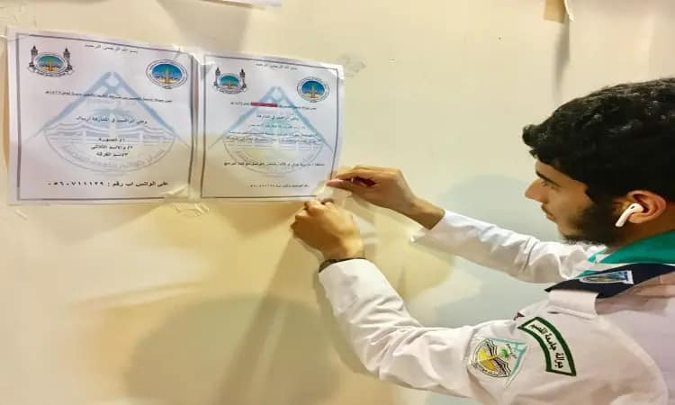 Wearing a Saudi shemagh and a Saudi t-shirt that gives him a touch of dignity, a young man is dressed in the official scout uniform of the university's Jawalat. He is pinning a modern paper notice with the slogans of serving the pilgrims and Arabic texts next to another notice, while the sleeve of his uniform is successfully adorned with scout badges and badges, and he is wearing a headset to complete his tasks efficiently and scheduled.