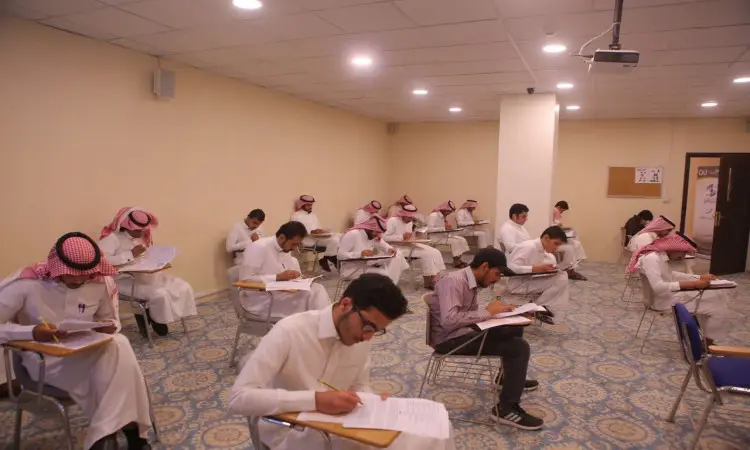 يجلس مجموعة من الطلاب، يخصص معظمهم الغترة أو الشماغ السعودي مع العقال والبشت السعودي التقليدي، متوزعين بمسافات مناسبة داخل قاعة الدخول في القصيم. استمتع كل طالب بأداء اختبار اللغة الإنجليزية على طاولة منفردة أمامه، بينما ستشاهدها أرضها المزخرفة وجدرانها ذات اللون الكريمي.