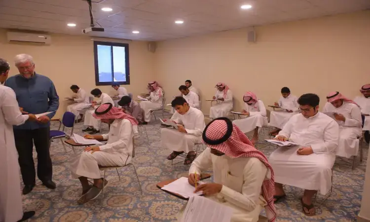 قاعة بها في جامعة القصيم يتواجد التلاميذ الزي السعودي الرسمي المكون من غترة أو شماغ سعودي، عقال، وبعضهم يرتدي البشت السعودي، وهم يؤدون اختبار إتقان اللغة الإنجليزية. اتصل برجل القاعة يتبادلان الحديث. تتميز الغرفة بأرضياتها المبلطة وجدرانها ذات اللون الكريمي ونافذة تسمح بدخول الضوء الطبيعي.