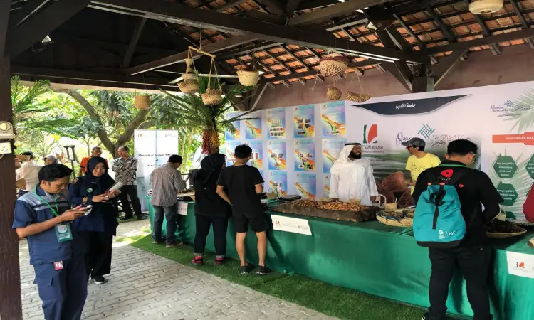 زار الزوار جناحاً في السوق المفتوح تحت مظلة خشبية ضمن فعاليات معرض النخلة في جاكرتا، حيث تم عرض الباعة ويبيعون التمور. للموظفين الصغار وهم ينتمون الغتة أو الشماغ والعقال والبشت السعودي، ويتفاعلون مع الباعة أو يقومون بتفقد هواتفهم. وتبرز في خلفية الرسوم المتحركة الدعائية التي تحمل شعارات جامعة القصيم، مما يضفي طابعًا على المعرضة ويعكس الهوية الثقافية السعودية.