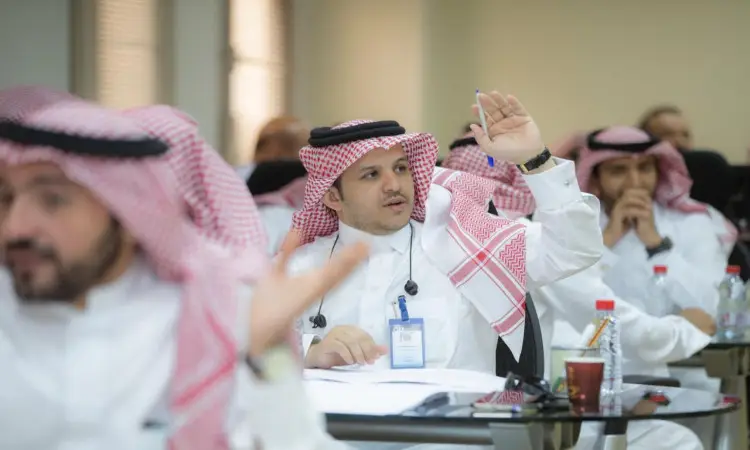 رفع أحد الرجال مرتديًا الغترة والعقال والبشت السعودي باليد خلال برنامج تدريبي في جامعة القصيم، بينما كان أعضاء هيئة التدريس يحيطون به. وقد جلس عدد من الحاضرين على الطاولات أمامهم وأوراق الشجر والمشروبات وبطاقات تعريفية ظاهرة.