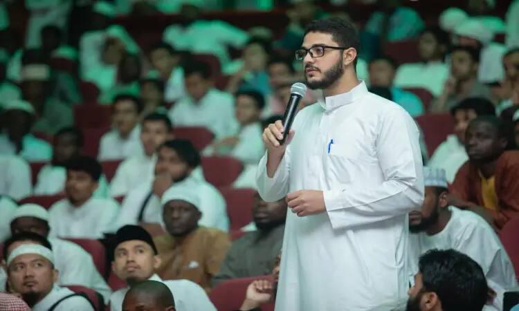 رجل القط مرتدياً غترة سعودية وعقالاً، وقد أكسى بالمشت السعودي فوق ثوبه الأبيض، وهو يمسك بيده تلقائياً ويتحدث أمام الحضور في قاعة محاضرات جامعة القصيم خلال لقاء مفتوح. يعتصمون في القاعة عدد كبير من المشاركين، وغالبيتهم من الرجال، يصغون باهتمام لما يطرح من نقاشات.