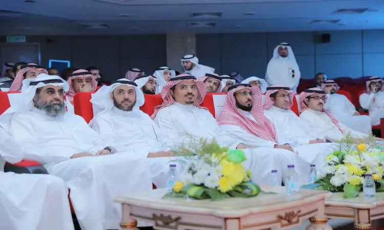 مجموعة من الرجال مرتدين الغترة أو الشماغ والعقال، بالإضافة إلى البشت السعودي، على كراسي حمراء في إحدى القاعات خلال فعالية يوم الجودة في جامعة القصيم. ووضعت على الطاولات أمامهم ترتيبات زهور وزجاجات صباحية احترام التنظيم الراقي للمناسبة. سجل في الخلفية من الحضور، بما في ذلك مدير الجامعة، وذلك في وسط المجتمع وإضاءة جيدة على أهمية الأهمية.