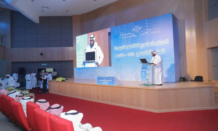 رجل مرتدياً الغترة السعودية والعقال والبشت السعودي، يكفي أنه مدير الجامعة، على المنصة مخاطباً يحضر فعاليات يوم الجودة في القصيم. يجلس الضيوف في الجزء السفلي من الشاشة الخلفية بينما يتم عرض الجزء الخلفي من الشاشة الرئيسية من النافذة الخلفية والفعاليات.