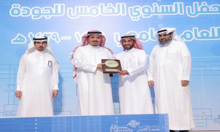 وقف الرجال اربعة الغترة أو الشماغ والعقال والبشت السعودي على منصة تكريم يوم الجودة في جامعة القصيم. يعمل رجلان في الوسط درع التكريم، ويبتسمان، بينما يتواصل مع مدير الجامعة ويضيف آخر على المشجعين. تواجد خلفهم لوحة زرقاء تحمل عبارات باللغة العربية.