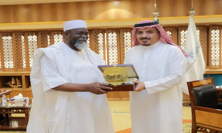 رجل الاتصال في مكان مغلق، يتبادلان الابتسامات أثناء حملهما درعاً ذهبياً. ارتدى سفير غانا زياً أفريقياً تقليدياً أبيض اللون مع قبعة قديمة، بينما ظهر مدير جامعة القصيم مرتدياً ثوباً سعودياً أبيض مع شماغ سعودي وعقال، وقد أتم إطلالته بارتداء البشت السعودي الرسمي.