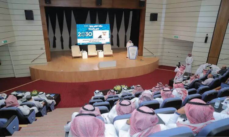 ظهير يرتدي ملابس سعودية تقليدية يشمل الغترة، الشماغ، العقال، والبشت السعودي، مخاطباً الحضور خلال الجامعة الفعالة بـ اليوم العالمي للمعلم. وقد ضمت منصة ثلاث كراسٍ مجانية وشاشة يُعرض عليها نص عربي وشعار 2030. جلسة معلمو المنطقة والضيوف في مواجهة المنصة أثناء الفعالية.