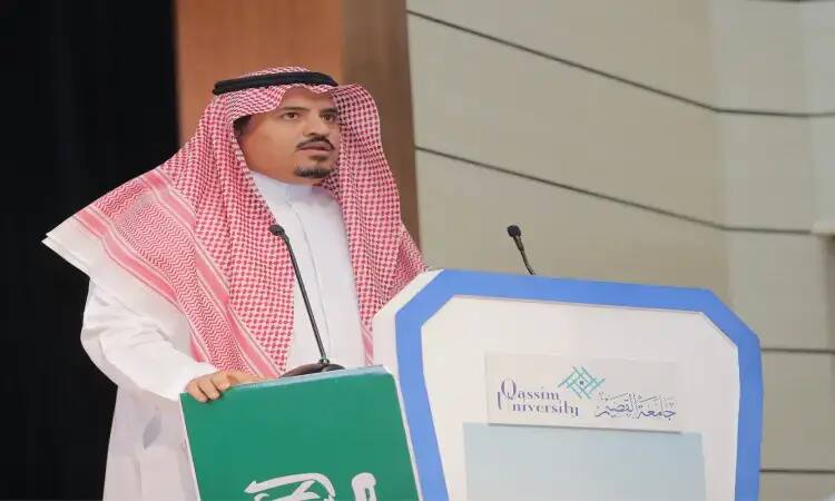 رجل مرتدياً غترة سعوديةقالاً وبشتاً تقليدياً وهو يتحدث من على منصة تحمل شعار جامعة القصيم، وذلك من خلال فعالية الجامعة بمناسبة اليوم العالمي للمعلم. وقد وقف في قاعة داخلية، ممسكًا لوحة خضراء كُتب عليها نص باللغة العربية، وخلفه جدار بلون وستارة مجسمة.