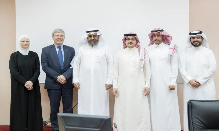 ستة أفراد جنبًا إلى جنب أمام شاشة سوداء في كلية الصيد في القصيم، وهم خمسة رجال الغترة أو الشماغ والعقال والبشت السعودي إلتزام السعودية، وسيدة ترتدي ملابس بيضاء. وقد التقطت لهم هذه الصورة الجماعية في مكان مغلق داخل الكلية.