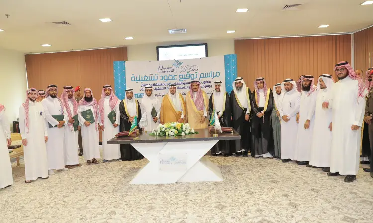 A group of men wearing the Ghutra or Shemagh and Eqal, and the Saudi Bisht, gather for a group photo during a contract signing ceremony at Qassim University. At the center of the scene is a table decorated with flowers and documents, while behind them is a plaque.