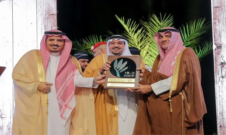 ثلاثة رجال أول الزي السعودي المكون من الغترة أو الشماغ والعقال والبشت السعودي، يبتسمون ويهتمون معاً لوحة جائزة جود الدولية على المسرح، احتفاءً بتكريمين، وذلك أمام خلفية تتزين بأشجار النخيل والأشجار الخشبية.
