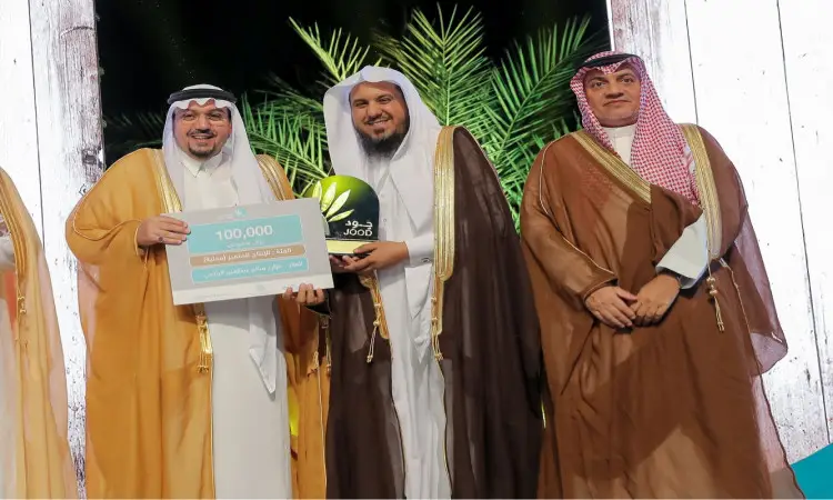 توقف ثلاثة رجال الغتة والشماع والعقال والبشت السعودي على تيموثاوس خلال أولمبياد. عصر الرجل الواقف إلى إيفان شيكاً 100,000 ريال سعودي، بينما يتوسطهم ممسك و جود الدولية. تعلو الابتسامة وجوههم وخلفهم نخلة وصورة صاحب السمو منطقة القصيم في مشهد رسمي يتبعق بمكانة جامعة القصيم.