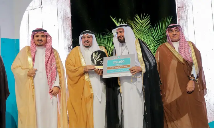 لأربعة رجال مرتدين الغترة أو الشماغ السعودي مع العقال والبشت السعودي على خشبة المسرح الفائزون خلال الاحتفالات. عصر أحدهم بينما يستعرض الآخر بشكل دقيق يبلغ مجموعها 250,000 ريال خلال جائزة جود الدولية، للاستهلاك في نخلة وقوس زخرفي، في نظمها جامعة القصيم.