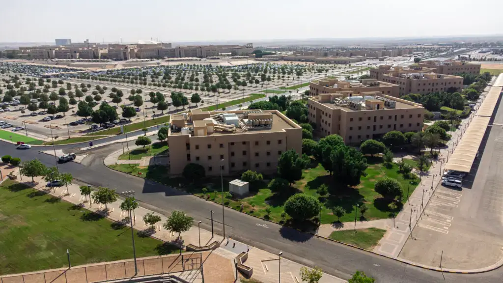 صورة طيران تركية لجامعة القصيم الخضراء عدداً من المباني ذات اللون المخصص لمواقف السيارات، تتخللها شوارع خضراء بالأشجار ومساحات تحت السماء صافية، بما في ذلك يبرز مخصصة للسكن والترفيه.