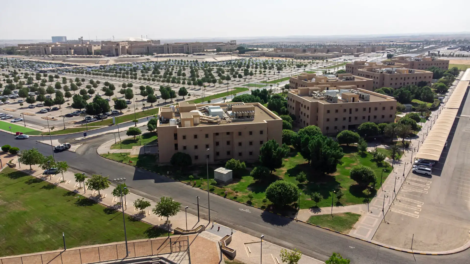 صورة طيران تركية لجامعة القصيم الخضراء عدداً من المباني ذات اللون المخصص لمواقف السيارات، تتخللها شوارع خضراء بالأشجار ومساحات تحت السماء صافية، بما في ذلك يبرز مخصصة للسكن والترفيه.