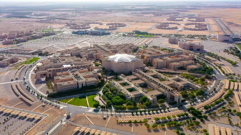 منظر جوي لجامعة القصيم يُبرز حرمها الجامعي الواسع والمتميز بالمباني والمساحات الخضراء والطرق الدائرية الحديثة، بالإضافة إلى مناطق مخصصة لكراسي البحثية، وكل ذلك ضمن بيئة تفاعلية للطبيعة النووية ومواقف السيارات، تحت مظلة سماء صافية.