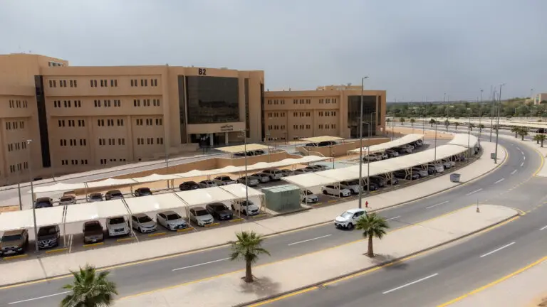 وتظهر في الصورة مبنى حديث وضخم ذو زجاج رملي عصرى ولا تحمل رمز (B2)، وتتميز بواجهته التي تتمتع بنوافذ زجاجية مع مدخل رئيسي واضح. أمام المبنى توزع مواقف سيارات مظللة تعج بالمركبات، وتحيط بعدد من سكان النخيل بالإضافة إلى الطريق الدائري ممهد. تغطيها السماء بعض الغيوم المتفرقة، مما يعكس الطابع العصري لسباق الجائزة في جامعة القصيم.