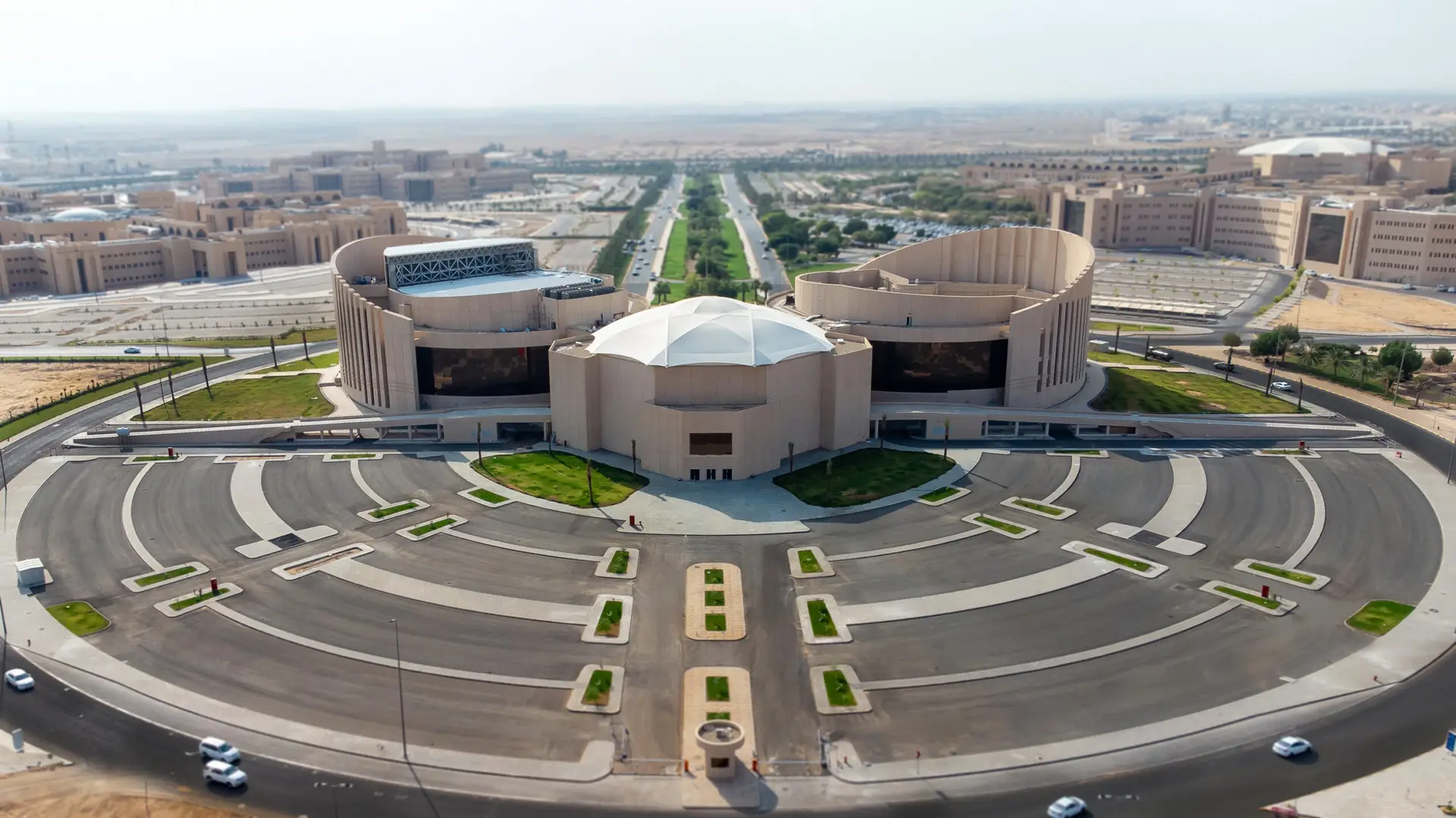 صورة جوية لمجمع مباني دائري ذي تصميم معماري منحني للسيارات وسقف على سطح المبنى فوق الطابق العلوي، يقع حديثًا داخل الحرم الجامعي في الحرم الجامعي في القصيم، وتحيط به وسائل منسقة ومناطق خضراء واسعة بالإضافة إلى مواقف.