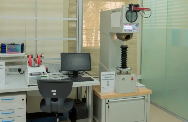 تضمين محطة العمل المخبرية المخصصة لتخصص الهندسة الميكانيكية في جامعة القصيم جهاز جهاز حاسب حديث، وكرسي أسود، وجهاز اختبار الصلادة الشامل من نوع من زايس (جهاز اختبار الصلابة العالمي من زايس)، بالإضافة إلى معدات مخبرية أخرى موضوعة على طاولة قريبة من النوافذ الزجاجية ذات الصلابة الستائر.