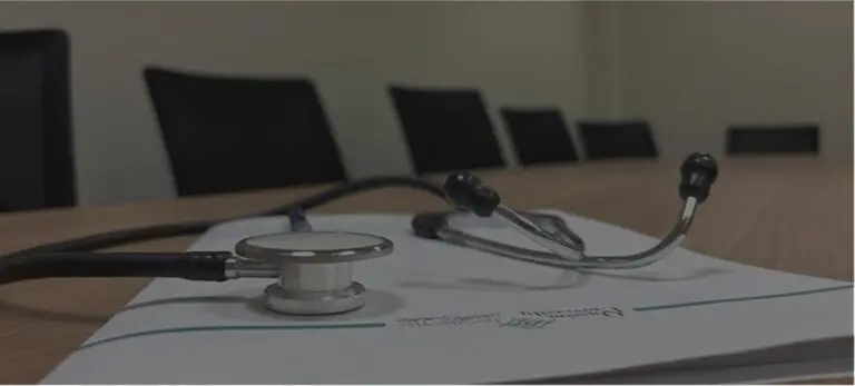 يرتفع سماعة الطبيب فوق مستند موضوع على طاولة اجتماعات خشبية، في مشهد يرمز إلى صحة المجتمع وتظهر في الخلفية كراساً سوداء خالية، ويُضفي على ذلك كله طابعاً رسمياً يناسب جامعة القصيم.