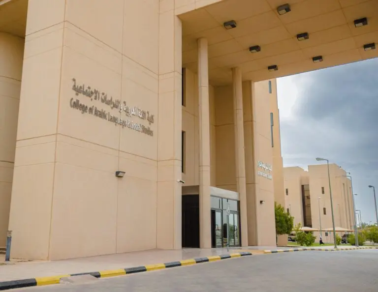 مدخل مبنى حديث أنيق، يتميز بأعمدة عتيقة وأبواب، مع لوحات تعريفية باللغتين العربية والإنجليزية تحمل اسم "كلية اللغة العربية والدراسات الاجتماعية (كلية العلوم والعلوم الإنسانية)". تسع بالمبنى ساحة مرصوفة بالأحجار والأشجار الخضراء، وتنظر إلى السماء ملبدة بالغيوم.