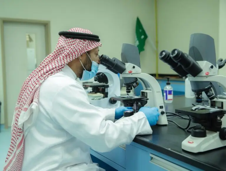 رجل يرتدي شماغاً أو غترة سعودية، يضاف إليها الكمامة والقفازات، يقوم بفحص المجهر المجهري في كلية الطب بمستشفى القصيم. لتحضير طاولة العمل مجاهر ومخبرية أخرى.