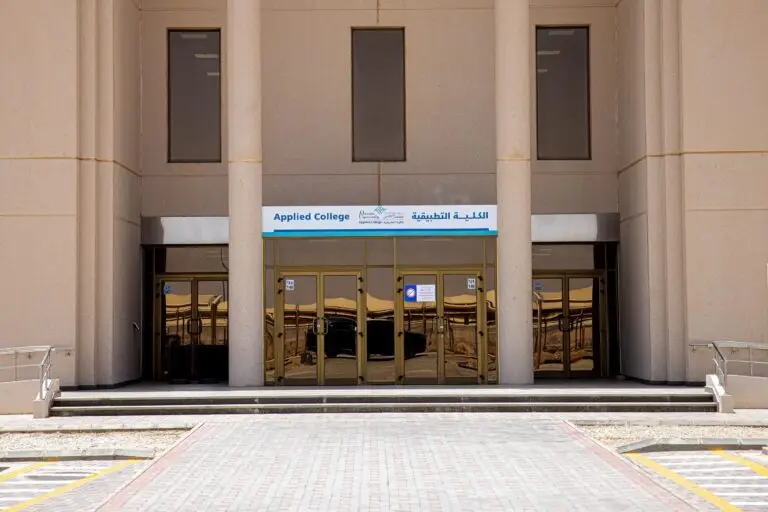 مدخل مبنى رمادي داكن بدون أبواب، تعلوه لوحة تحمل اسم "الكلية التطبيقية" باللغتين العربية والإنجليزية، ويستند المدخل إلى أربع أعمدة كبيرة. ويقود ممر مرصوف بالطوب إلى بوابة الدخول.