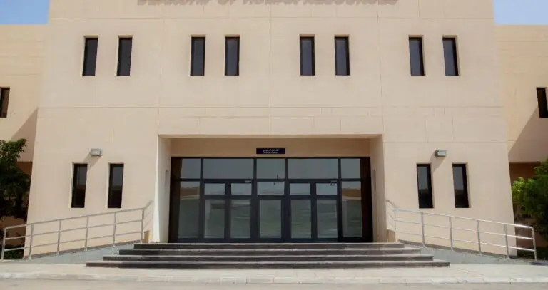 تُظهِر الصورة مدخل مبنى الإدارة العامة ذات اللون البيج، ويتكون من أبواب مزدوجة زجاجية تعلوها ست نوافذ ضيقة يحيط بالمدخل سلم عريض ومنحدران على الجانبين الوصول، وتحيط بهما أشجار صغيرة ومصابيح خارجية.