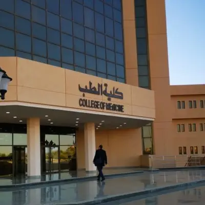 A man wearing a Saudi shemagh or a Saudi ghutra walks toward a modern entrance with large windows and pigeon-colored walls. A sign at the entrance reads "Faculty of Medicine" and "Department of Psychiatry" in both Arabic and English, with a sign that reads "Faculty of Medicine" and "Department of Psychiatry.