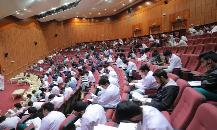 قاعة محاضرات واسعة في جامعة القصيم، يجلسون فيها طلبة كليات الطب على الأحمر وهم منهمكون في الكتابة، ما يوحي الذين يؤدون اختبار القياس المستوى. يظهر الطلاب بملابس تتناسب مع البيئة الجامعية، حيث يجتمع عدد من الطلاب الغترة أو الشماغ والعقال، ويضع بعض أعضاء هيئة التدريس البشت السعودي. يسود القاعده من الجديه والهدوء يعكس دلالة صحيحة وأجوبة الانضباط الأكاديميين في الجامعة.