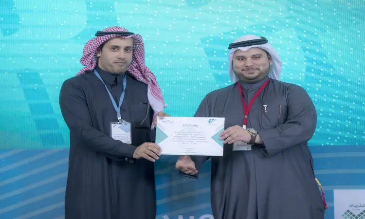 Two men dressed in Saudi uniforms, wearing the Ghutrah and Eqal and the Saudi Bisht, next to a blue background decorated with an old-fashioned pattern. Both are smiling as they compete in competitions at Qassim University.