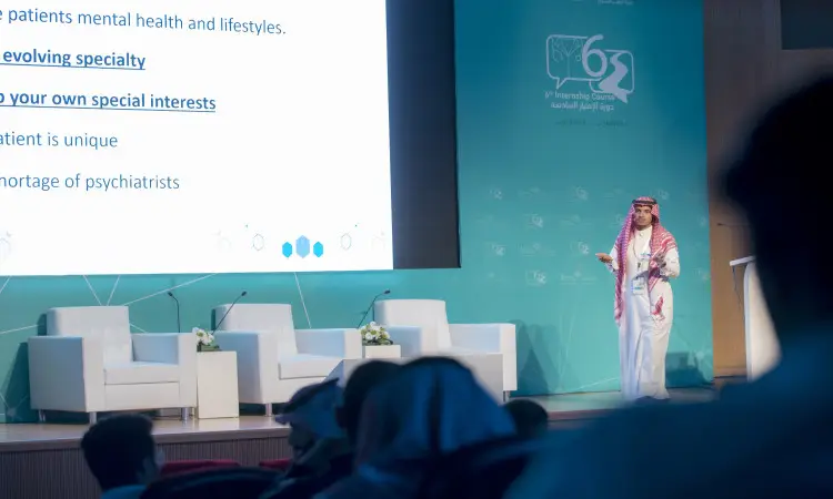 رجل يرتدي الزي الوطني السعودي الاثني عشر في غترة سعودية وقال وبشتدعى على منصة المؤتمر، المتحدث باسمه أمام الحضور، وبجواره شاشة ضخمة تعرض نصوصًا حول علم الطب النفسي. يتخصص في دورة الامتياز الخاصة بالأطباء المتميزين من كلية الطب في القصيم. استمعوا إلى الصفوف، استمعوا إلى كلمته الرسمية.