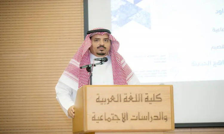 رجل القط مرتدياً غترة سعودية مع شماع وقال وبشت سعودي عند منصة الخطابة، أمام الحضور. تحتوي على شريحة عرض على النص باللغة العربية تمثل كلية اللغة العربية في جامعة القصيم. عاكس الضوء للمدرسة المدرسية أو المدرسة الرسمية المستوى.