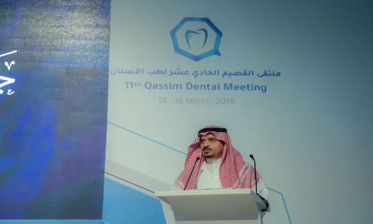 مدير الجامعة مرتدياً الزي السعودي التقليدي الامتداد في الغترة أو الشماغ والعقال والبشت السعودي، مشهوراً من على المنصة خلال فعاليات ملتقى القصيم لطب الأسنان 2019. للاستهلاك خلفه تفاصيل الملتقى وشعاره المتعلق بطب الأسنان على الخلفية الرسمية للحدث.